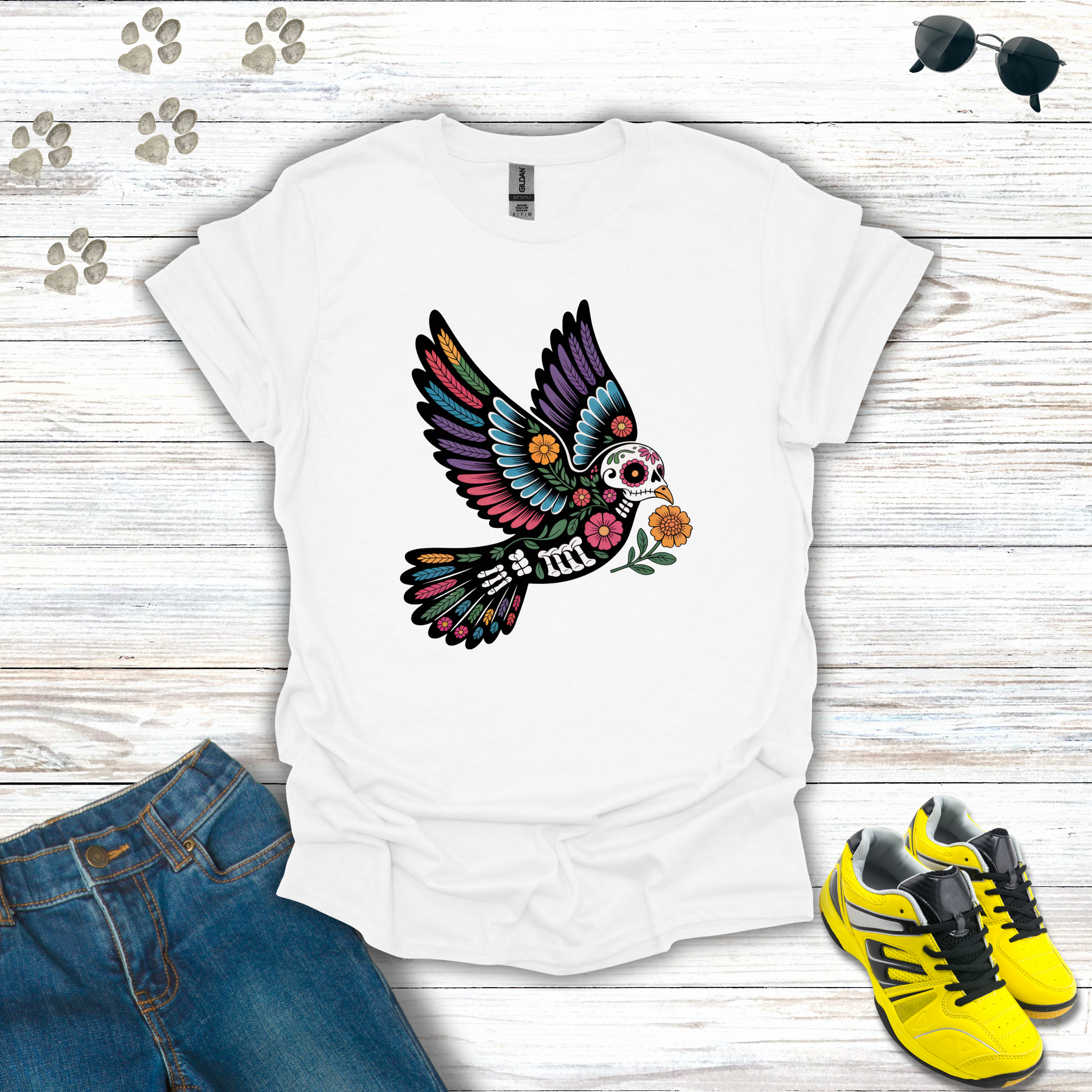 White t-shirt with colorful skeleton dove design on a wooden surface with jeans and sneakers.