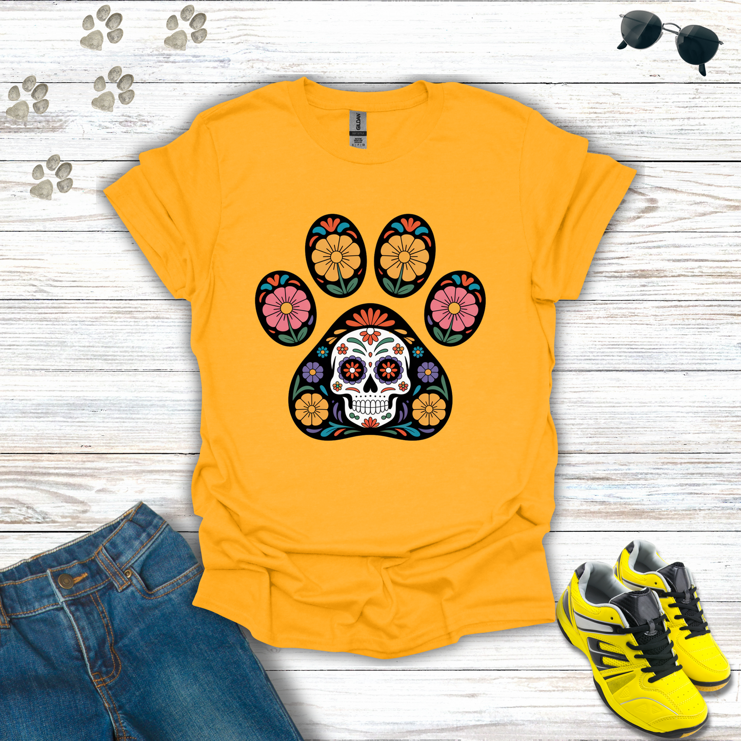 Gold t-shirt with a colorful paw print design featuring a skull, placed on a wooden surface with jeans and sneakers.
